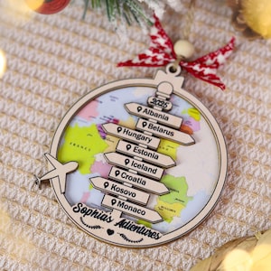 May include: Wooden Christmas ornament with a world map design and directional signs pointing to various countries. The ornament includes the text "Sophia's Adventures" and a small airplane cutout. It is decorated with a red and white ribbon.