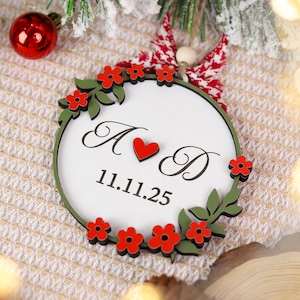 May include: A round, white Christmas ornament with a green border, red flowers, and green leaves. The ornament features the letters "A" and "D" with a red heart, and the date "11.11.25". A red and white bow is at the top.