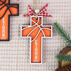 May include: Wooden cross ornament with a basketball design in orange and white. The name "ETHAN" is printed vertically on the cross, along with the date "Baptized 02.17.25". The ornament is decorated with a red and white bow.