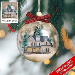 May include: A clear glass snow globe ornament with a red and black plaid ribbon bow. Inside the globe is a miniature white house with a gray roof and two small green trees. The globe is sitting on a green tree branch.