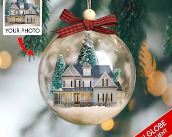 May include: A clear glass snow globe ornament with a red and black plaid ribbon bow. Inside the globe is a miniature white house with a gray roof and two small green trees. The globe is sitting on a green tree branch.
