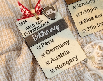 May include: Wooden travel-themed ornament with the name "Bethany" and the text "Road Trip Extravaganza 2024." Features a shield with a camper graphic and check boxes for countries like Peru, Germany, Austria, and Hungary. Includes a red and white ribbon.