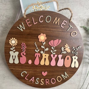 May include: A wooden round sign with the text "Welcome to Ms. Taylor Classroom" in colorful letters. The sign is decorated with flowers and vases in pink, orange, and green.