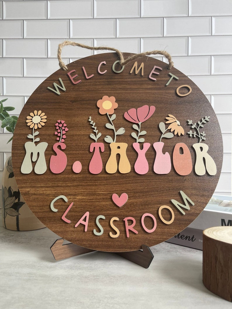 May include: Welcome to Ms. Taylor's Classroom sign with a brown wooden background and colorful letters. The sign features a heart and flowers in pots.