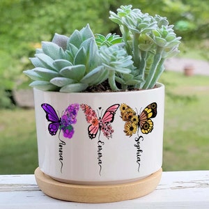 May include: White ceramic planter with three butterflies, each with a different floral design and a name underneath: Anna, Emma, Sophia. The planter has a wooden base and is filled with green succulents.