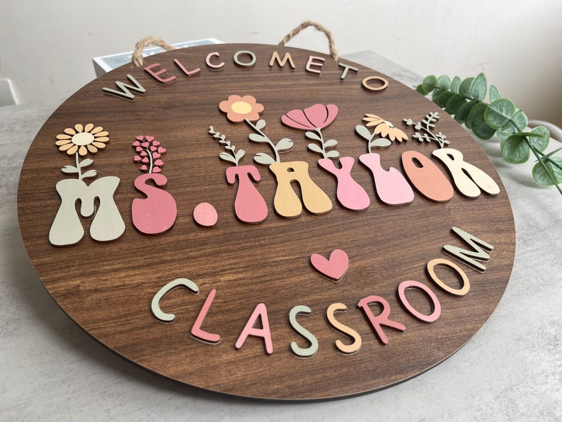 May include: A round wooden sign with the text "Welcome to Ms. Taylor's Classroom" in colorful letters. The letters are decorated with flowers and hearts.