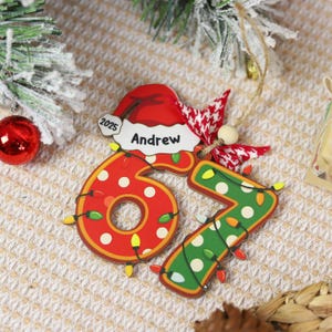 May include: A personalized Christmas ornament featuring the number "67" with festive lights. The number "6" is red with white polka dots, and the number "7" is green with white polka dots. A Santa hat with the name "Andrew" and the year "2025" is also included.