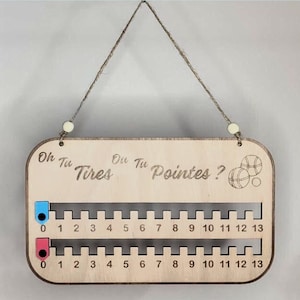 Peut inclure: Panneau de jeu en bois avec le texte "Oh Tu Tires Ou Tu Pointes ?" et deux pistes numérotées de 0 à 13. Le tableau a une ficelle pour l'accrocher et comporte des marqueurs bleus et roses. Le tableau de jeu est de couleur bois clair.