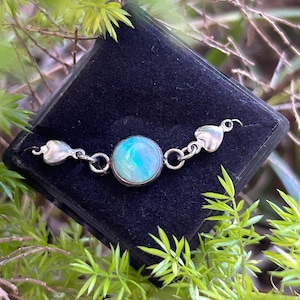 May include: A silver bracelet featuring a round, iridescent blue-green opal gemstone. The bracelet has heart-shaped details at each end and is presented on a black velvet surface, framed by green leaves.