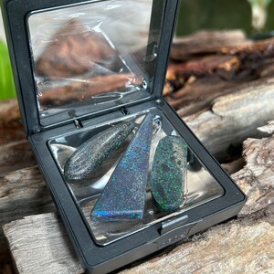 May include: Three iridescent black opal stones in a black square box with a clear lid. The stones are different shapes and sizes, with a teardrop, a triangle, and a rectangle.