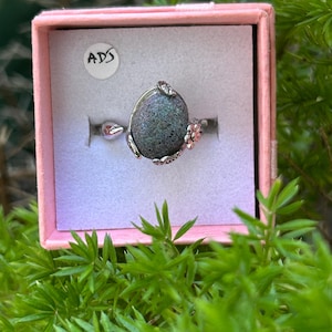 Op de afbeelding: Een zilveren ring met een grote, ovale, donkergrijze steen. De steen is omgeven door zilveren accenten en kleine roze bloemen. De ring wordt gepresenteerd in een roze doos met een wit interieur en een sticker met "ADS".
