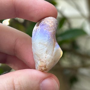 Puede incluir: Una piedra preciosa de ópalo natural, sostenida entre los dedos, muestra una mezcla de colores. La piedra tiene una superficie rugosa y texturizada con áreas blancas, beige y tonos iridiscentes azules y amarillos. El ópalo tiene forma irregular.