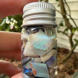 Può includere: Un piccolo barattolo di vetro trasparente con un tappo a vite color argento, riempito con una collezione di frammenti di opale irregolari e multicolori. Le pietre mostrano bagliori iridescenti di blu, verde e bianco, visibili attraverso il vetro.