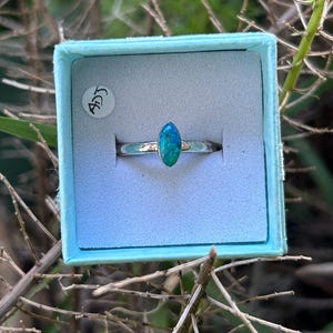 May include: A silver ring featuring an oval, blue-green gemstone, presented in a light blue jewellery box. The ring is centrally placed, with the gemstone reflecting light. The box has a white sticker with the letters "ADJ".
