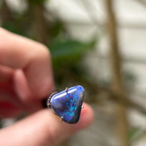 May include: A triangular-shaped opal ring with a dark blue base color and vibrant blue and green flashes. The gemstone is held in place by three silver prongs. The ring is a unique piece of jewelry.