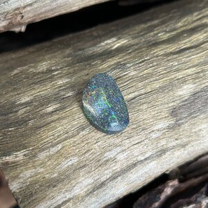 Puede incluir: Una piedra preciosa de ópalo ovalada de color gris oscuro con destellos iridiscentes verdes, azules y negros. El ópalo está sobre un trozo de madera desgastada.