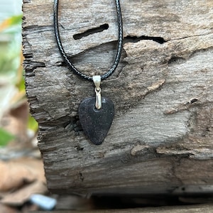 Puede incluir: Un collar de cordón de cuero negro con un colgante de corazón marrón oscuro. El colgante tiene una anilla de plata y un pequeño agujero en la parte superior.