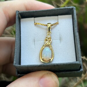 Puede incluir: Un collar con colgante dorado con una piedra de &oacute;palo ovalada e iridiscente. El colgante tiene forma de l&aacute;grima, con un peque&ntilde;o detalle de diamante en la parte superior. El collar se muestra en una peque&ntilde;a caja de joyer&iacute;a gris.