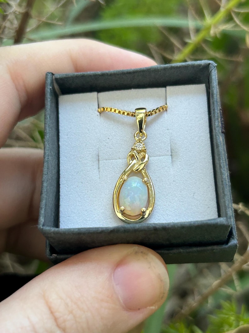 Puede incluir: Un collar con colgante dorado con una piedra de &oacute;palo ovalada e iridiscente. El colgante tiene forma de l&aacute;grima, con un peque&ntilde;o detalle de diamante en la parte superior. El collar se muestra en una peque&ntilde;a caja de joyer&iacute;a gris.