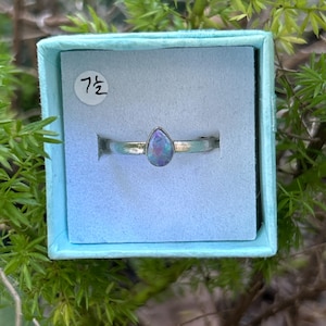 May include: A silver ring with a pear-shaped, iridescent opal gemstone. The ring is displayed in a light blue box with the number "7" printed on a white circle. The background features green foliage.