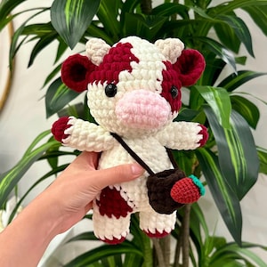 Pode incluir: Uma vaca de pelúcia de crochê com manchas brancas e vermelhas. A vaca tem um nariz rosa e olhos pretos. A vaca está segurando uma bolsa de crochê marrom com um morango vermelho.