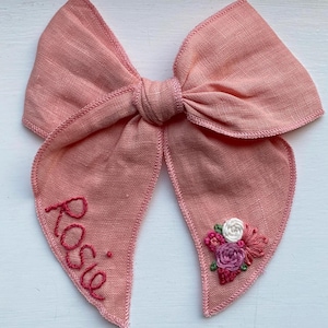 May include: A pink fabric bow with the name "Rosie" embroidered on one side and a pink, white and green floral design embroidered on the other side.
