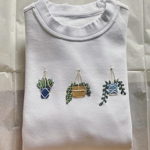 May include: White sweatshirt with three embroidered hanging plants. The plants are in different colored pots, one blue, one yellow, and one blue and white.
