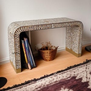 May include: A decorative console table with a curved design. The table features a white and brown leaf pattern with gold trim. A woven basket and a stack of magazines are visible beneath the table. The table is placed on a wooden floor and a patterned rug.