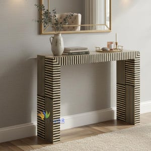 Puede incluir: Una consola decorativa con un patrón de rayas blancas y negras. La mesa tiene una parte superior rectangular y dos patas con un diseño geométrico. Un espejo, un jarrón, libros y velas están sobre la mesa. La pared es de color gris claro y el suelo tiene una alfombra blanca.