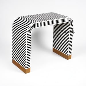 Puede incluir: Una mesa consola tipo cascada con un patrón de rayas blancas y negras en toda su superficie. La mesa presenta una parte superior y laterales curvos, con bases de metal en tono dorado sólido en la parte inferior de cada pata para mayor estabilidad.