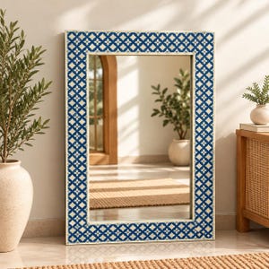 May include: A rectangular wall mirror featuring a wide frame with a deep blue background and a repeating white geometric floral pattern. The mirror glass is clear and reflective, showing a room interior with a potted plant and a wooden archway.
