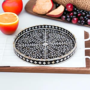 May include: A round black tray with a white floral design made of mother of pearl. The tray has a decorative border with a diamond pattern.