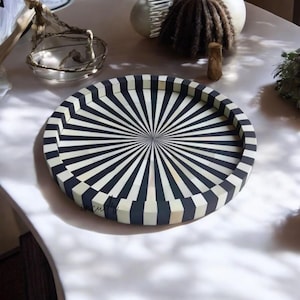 May include: A circular decorative tray featuring a black and white striped design, with lines emanating from the centre. The tray has a raised edge and is made of a solid material. The tray is on a white surface.