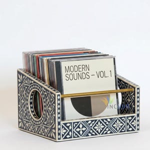May include: A rectangular storage rack featuring a black and white geometric inlay pattern with a circular cutout on the side panels. A horizontal brass-toned metal bar secures a collection of CD cases, including one labeled Modern Sounds - Vol. 1.