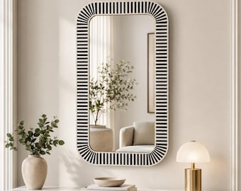 May include: A rectangular wall mirror with rounded corners, featuring a frame decorated with a repeating pattern of vertical black and white stripes. The mirror surface reflects a room interior with a plant and furniture, showing clear glass.