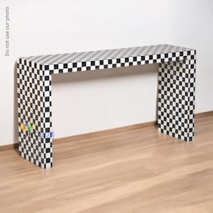 Puede incluir: Una consola a cuadros en blanco y negro. La mesa tiene una parte superior rectangular y dos patas, con un patrón de tablero de ajedrez. La mesa está hecha de un material sólido y es adecuada para un pasillo o sala de estar.
