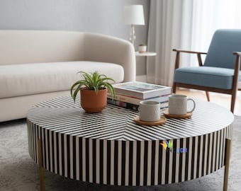 Op de afbeelding: Een ronde salontafel met een zwart-wit gestreept patroon op het blad en de zijkanten, ondersteund door dunne goudkleurige metalen poten. Op de tafel staan een kleine terracotta plantenpot, een stapel boeken en twee gespikkelde mokken op onderzetters.