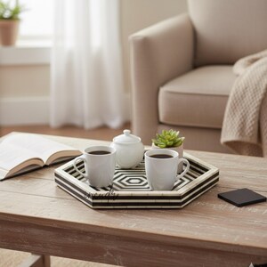 May include: A decorative hexagonal tray with black and white geometric patterns holds two white mugs filled with coffee. A white sugar bowl and a small succulent plant sit on the tray. A book and a coaster are also on the wooden coffee table.