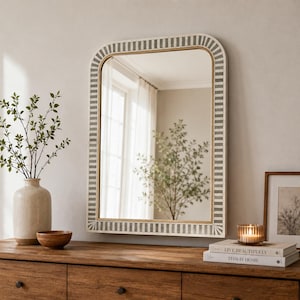 Puede incluir: Un espejo de pared rectangular con esquinas superiores redondeadas, que presenta un marco con un patrón repetitivo de rayas verticales grises y blancas. El espejo refleja una habitación luminosa con una ventana y un árbol, montado en una pared neutra.