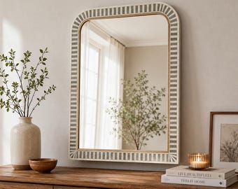 May include: A rectangular wall mirror with rounded top corners, featuring a frame with a repeating pattern of vertical grey and white stripes. The mirror reflects a bright room with a window and a tree, and it is mounted on a neutral wall.
