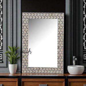 Könnte beinhalten: Rechteckiger Spiegel mit dekorativem Rahmen mit geometrischem Muster in neutralen Tönen. Der Spiegel ist auf einem dunklen Holzschrank montiert, mit einer kleinen Pflanze in einem weißen Topf links. Das Wort "render" ist auf dem Spiegel geschrieben.
