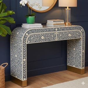 May include: A console table featuring a dark blue surface with intricate white floral and geometric bone inlay patterns. The table has two drawers, a curved waterfall silhouette, and rectangular gold-toned metal bases at the bottom of each side.