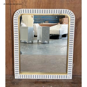 May include: A large rectangular mirror with a white and gray striped frame. The frame is made of bone inlay and has a gold trim. The mirror is reflecting a room with furniture.
