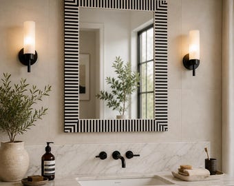 May include: A rectangular wall mirror featuring a bold black and white striped frame with mitered corners. The mirror is mounted on a light-colored wall between two cylindrical wall sconces with frosted glass shades and black metal hardware.
