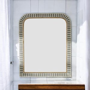 May include: Rectangular wall mirror with a rounded top. The frame features a repeating pattern of gray and white stripes. The mirror is set against a white tiled wall, with a wooden cabinet visible below.