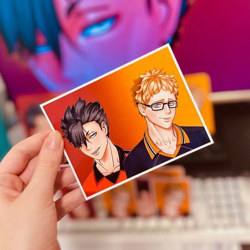 Puede incluir: Una impresi&oacute;n art&iacute;stica rectangular con dos personajes de estilo anime sobre un fondo degradado naranja y rojo. Un personaje tiene cabello negro, el otro tiene cabello rubio y gafas. La impresi&oacute;n est&aacute; sostenida por una mano.