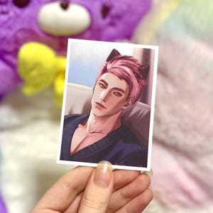 May include: A rectangular sticker featuring a digital illustration of a person with short pink hair and dark cat ears. The person has amber eyes and wears a dark blue V-neck top. The art style is clean and detailed, showing the head and shoulders.
