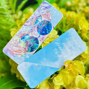 May include: Two bookmarks, one with a blue sky and clouds and the text "I have loved and dreams too", and the other with a holographic image of a person with blue eyes and a holographic design.