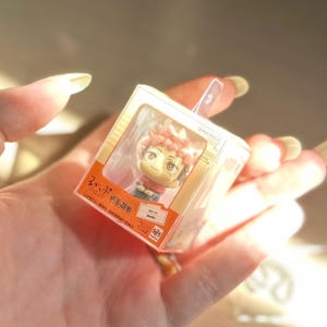 May include: A small, clear plastic display box containing a miniature anime-style figurine with spiky pink hair and brown eyes. The packaging features orange accents, Japanese text, and the Bandai Namco logo, designed for collectible display purposes.