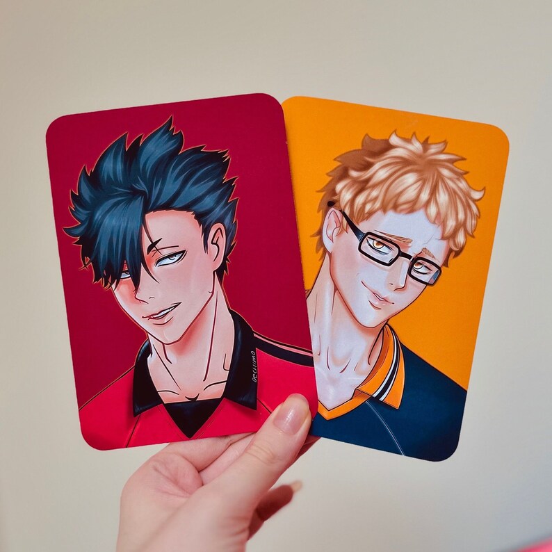 Puede incluir: Dos impresiones art&iacute;sticas rectangulares con ilustraciones de estilo anime. Una tiene un fondo rojo con un personaje de pelo azul oscuro. La otra tiene un fondo naranja con un personaje de pelo rubio y gafas. Las impresiones son sostenidas por una mano.